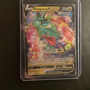 Rayquaza V Pokémonkort - Rayquaza V är ett samlarkort från Pokémon TCG med färgstark design och holografisk yta. Kortet har motiv av Rayquaza i grönt och rött med guldiga detaljer och är inneslutet i ett genomskinligt plastfodral. Perfekt för dig som gillar Pokémon och samlarkort.