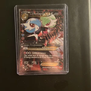 Pokémonkort Mega Gardevoir EX - Säljer ett samlarkort från Pokémon, Mega Gardevoir EX. Kortet har en färgstark design med Gardevoir i blått, grönt och rött mot en glittrig bakgrund. Kortet är inplastat i ett genomskinligt plastfodral för extra skydd.