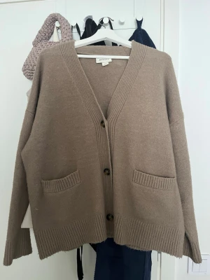 Beige kofta med fickor  - Mysig beige kofta med v-ringning, knäppning framtill och två fickor. Koftan har ribbade muddar vid ärmslut och nederkant samt en avslappnad passform. Perfekt för lager på lager och enkel att styla. Den är i xxs men oversized så passar mig som har s