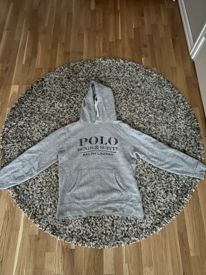 Grå hoodie från Polo Ralph Lauren - Säljer en grå hoodie från Polo Ralph Lauren med trycket 'POLO DENIM & SUPPLY' på bröstet. Hoodien har en klassisk känguruficka framtill och justerbar huva med snören. Tillverkad i mjukt bomullsmaterial för skön känsla. Perfekt för en avslappnad och stilren look.