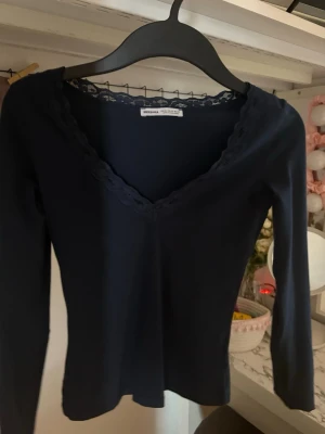 Mörkblå långärmad topp med spets - Mörkblå långärmad topp från Bershka med v-ringning och dekorativ spetskant runt halsen. Tunn och stretchig modell som sitter snyggt på kroppen. Perfekt att styla med jeans eller kjol för en trendig look.