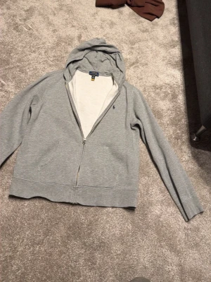 Grå zip hoodie från Polo Ralph Lauren - Snygg grå hoodie med dragkedja från Polo Ralph Lauren. Klassisk design med liten mörkblå logga på bröstet, huva och långa ärmar. Tillverkad i mjukt bomullsmaterial som är skönt att ha på sig. Perfekt för en avslappnad och stilren look. Det är storlek XL i barn storlek 100% äkta