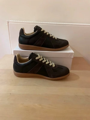 Maison margielas  - Snygga svarta sneakers med mörkbruna mockadetaljer och beige snören. Skorna har en klassisk låg profil och gummisula i ljusbrunt. Insidan är ljusbeige och designen är stilren, perfekt till jeans eller streetwear. Materialmix av mocka och läderimitation.