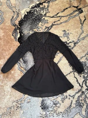Svart kort klänning med spetsdetaljer - Otrlolig vacker svart kort klänning med långa ärmar och v-ringning. Överdelen har fina spetsdetaljer och broderat mönster, medan ”kjolen” är enkel och flowy. Klänningen har dragkedja i ryggen och markerad midja för en snygg siluett. Passar perfekt för en sommardag ute med vänner eller på bröllop. Anledning till att jag säljer denna är på grund av att den är för kort på mig. 