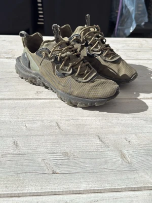 Nike React Element 55 sneakers - Säljer ett par Nike React Element 55 sneakers i olivgrönt och svart. Skorna har en sportig design med robust sula, mesh- och textilmaterial samt reflekterande detaljer. Perfekta för dig som gillar streetwear och outdoor-vibe.