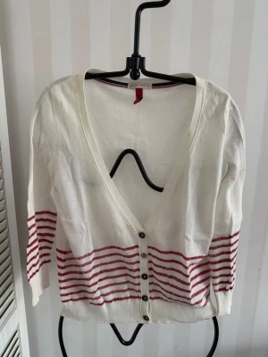 Vit kofta med röda ränder QS  - Vit tunn kofta från H&M med röda horisontella ränder nedtill och på ärmarna. Djup V-ringning och knäppning framtill med små knappar. Lätt och luftig modell, perfekt för lager på lager. Materialet känns mjukt och stretchigt.