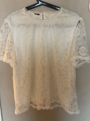 Vit spetsblus med kort ärm - Säljer en superfin vit blus i spets med korta ärmar och rund hals. Blusen har ett romantiskt mönster och är perfekt för dig som gillar en feminin stil. Passar till både jeans och kjol. Spetsdetaljerna ger en extra lyxig känsla.