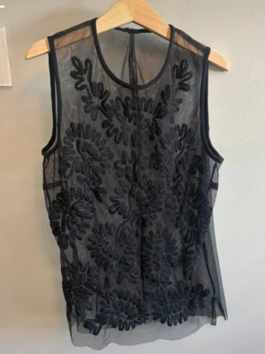 Svart broderad ärmlös topp - Snygg svart ärmlös topp i transparent mesh med broderade blommönster framtill. 