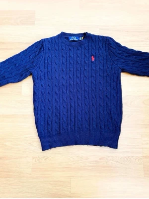 Mörkblå kabelstickad tröja Polo - Kabelstickad mörkblå tröja från Polo Ralph Lauren med rund halsringning och röd broderad logga på bröstet. Tröjan har ribbade muddar vid ärmslut och nederkant, perfekt för dig som gillar klassisk och stilren design.