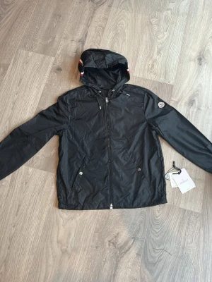 Svart vindjacka från Moncler - Snygg svart vindjacka från Moncler med huva och dragkedja framtill. Jackan har två fickor med tryckknappar och Moncler-logga på ärmen. Materialet är tunt och lätt, perfekt för blåsiga dagar. Justerbar huva med snören och diskreta detaljer. Storlek 2 vilket är M men funkar bra till S också då den är mellan dem två storlekarna. 