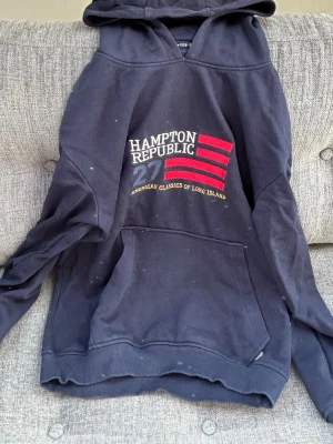 Marinblå hoodie från Hampton Republic - Snygg marinblå hoodie från Hampton Republic med tryck framtill i vitt, rött och gult. Klassisk känguruficka och ribbade muddar. Stor huva och mjukt material, perfekt för en avslappnad stil. Texten 'American Classics of Long Island' ger en cool amerikansk vibe.