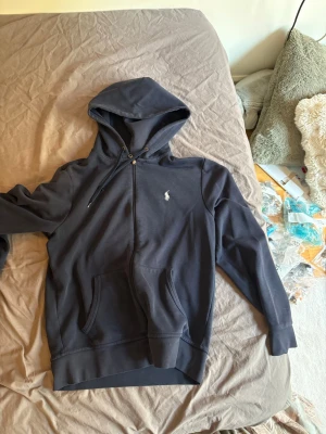 Mörkblå hoodie från Polo Ralph Lauren - Snygg mörkblå hoodie från Polo Ralph Lauren med dragkedja framtill och klassisk logga broderad på bröstet. Hoodien har fickor på magen, justerbar huva med snören och är tillverkad i mjukt bomullsmaterial. Perfekt för en avslappnad och stilren look.