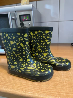 Svarta gummistövlar med gult mönster - Säljer ett par svarta gummistövlar med gult stenmönster. Stövlarna har en klassisk siluett, rund tå och dragflik bak för enkel påtagning. Tillverkade i slitstarkt gummi som håller tätt mot regn och slask.