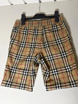 Beige rutiga shorts från Burberry - Säljer ett par klassiska beige shorts från Burberry med det ikoniska rutiga mönstret i svart, vitt och rött. Shortsen har elastisk midja och sidofickor, perfekta för en avslappnad och stilsäker look. KVITTO FINNS!! Pris kan diskuteras.