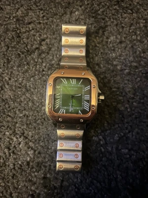  Santos klocka i silver - Snygg  Santos klocka med fyrkantig urtavla i grönt och romerska siffror. Armbandet är i silverfärgad metall med guldfärgade skruvdetaljer. Klassisk och lyxig design som sticker ut.