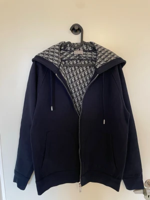 Marinblå hoodie från Dior - Snygg marinblå hoodie från Dior med dragkedja och två stora fickor framtill. Insidan av huvan har ett ikoniskt Dior-mönster i svart och vitt. Tillverkad i mjukt material och har justerbara snören i huvan. Perfekt för en trendig och avslappnad stil. Storlek M men passar även S. Säljer för 5000kr, priset är inte fast och kan förhandlas vid snabb affär 