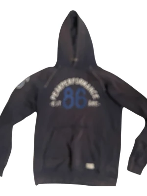 Mörkblå Åre hoddie - Snygg svart hoodie från Peak Performance med stor huva och tryck i vitt och blått på bröstet där det står 'Peak Performance 86 EST Åre'. Klassisk känguruficka framtill och dragsko i huvan. Perfekt för en avslappnad streetstyle-look. 2 defekter på bild 4 och 5. Dåliga bild pga kameran är sönder! 