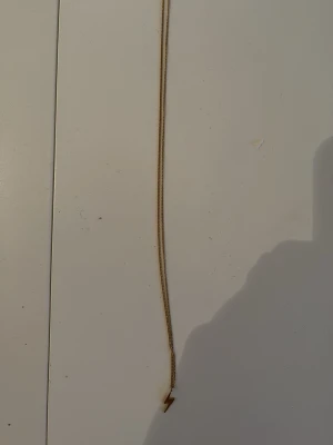 Guldhalsband med blixtberlock - Snyggt halsband i guldfärgad metall med en liten blixtformad berlock. Kedjan är tunn och stilren, perfekt för att addera en cool detalj till din outfit. Ingen synlig märkning på halsbandet.