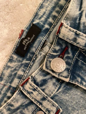 True Religion Bella jeans med slitningar - Säljer ett par ljusblå Bella jeans från True Religion med cool acid wash och röda kontrastsömmar. Jeansen har klassisk femficksmodell, silverfärgade knappar och slitna detaljer för en edgy look. Perfekta för dig som gillar streetstyle och vill sticka ut. Becca 