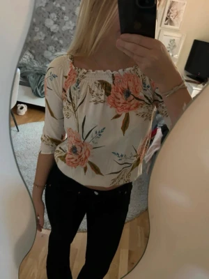 Blommig offshoulder blus i vitt - Supersöt blus med offshoulder och trekvartsärm. Vit botten med stora blommiga mönster i rosa och grönt. Smockad kant upptill och avslappnad passform. Perfekt för dig som gillar romantisk och trendig stil.