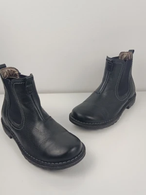 Ecco Chelsea Boots Svarta Strl 37 - Ett par svarta Chelsea boots med resårpaneler på sidorna för enkel på- och avtagning. Skorna har en robust sula med bra grepp och en klassisk design som passar till många olika tillfällen. Det finns en liten skada på sulan, titta sista bild. Jag tror att namnet av skorna är Bella.