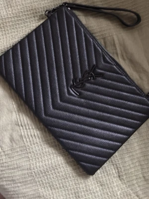 Svart kuvertväska - Snygg svart kuvertväska i quiltat läder imitation med logga framtill. Väskan har dragkedja upptill och en avtagbar handledsrem. Perfekt accessoar för att lyfta din outfit med en lyxig touch.