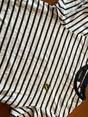 Randig t-shirt från Lyle & Scott - Säljer en vit och marinblå randig t-shirt från Lyle & Scott med klassisk gul logga på bröstet. T-shirten har korta ärmar och är gjord i mjuk bomull, perfekt för en avslappnad och stilren look.