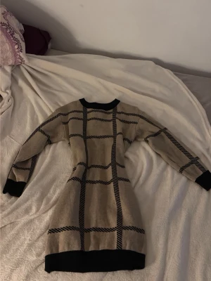 Beige och svart rutig stickad klänning - Säljer en beige stickad klänning med svart rutmönster och svarta muddar vid hals, ärmslut och nederkant. Klänningen har lång ärm och en kroppsnära passform som framhäver siluetten. Perfekt för dig som gillar stilrena och klassiska plagg.