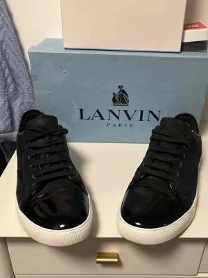 Svarta Lanvin sneakers med lackdetaljer - Snygga svarta sneakers från Lanvin med glansiga lackdetaljer på tån och klassisk vit sula. Skorna har svarta skosnören och en stilren design som passar till det mesta. Materialmix av läder och textil ger en exklusiv känsla.