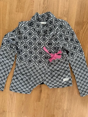 Odd Molly - Mönstrad kofta med rosa rosett - Unik svartvit kofta med grafiskt mönster och hög krage. Dekorerad med en rosa rosett på bröstet och har två fickor framtill. Perfekt för dig som gillar statement-plagg och vill sticka ut lite extra.