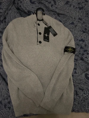 Grå stickad tröja från Stone Island - Snygg grå stickad tröja från Stone Island med hög krage och fyra svarta knappar framtill. Tröjan har ribbade muddar och den klassiska Stone Island-loggan på vänster ärm. Perfekt för dig som gillar stilrena och exklusiva plagg.