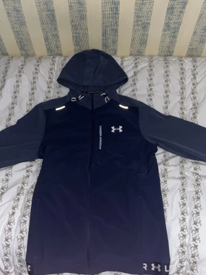 Mörkblå vindjacka från Under Armour - Snygg mörkblå vindjacka från Under Armour med grå ärmar och huva. Jackan har hel dragkedja, två sidofickor och reflekterande detaljer vid axlarna. Tillverkad i ett lätt och vattenavvisande material, perfekt för blåsiga dagar. Under Armour-logga på bröstet.