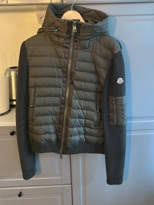 Olivgrön dunjacka från Moncler - Snygg olivgrön dunjacka från Moncler med stickade ärmar och huva. Jackan har quiltad front, dragkedja, ribbade muddar och en ficka med dragkedja på ärmen. Perfekt för kalla vinterdagar och har Moncler-logga på armen.