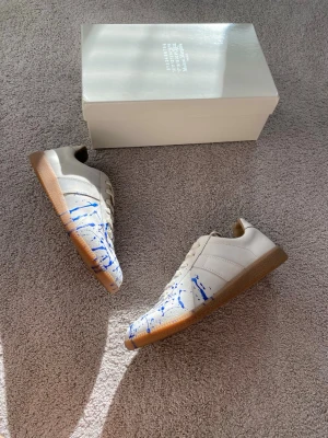 Maison Margiela Blue Paint Splatter Gats - Unika vita sneakers från Maison Margiela med blå färgstänk och klassisk gummisula. Skorna har lågt skaft, vita skosnören och är tillverkade i läder. Perfekt för dig som vill sticka ut med en kreativ och trendig look. True to size.