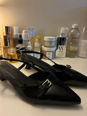 Svarta lackpumps med rem och låg klack - Säljer ett par svarta pumps i glansigt lackmaterial med spetsig tå och låg klack. Skorna har en smal rem med silverfärgat spänne över foten och öppen häl. Perfekta för dig som gillar en klassisk men trendig look.