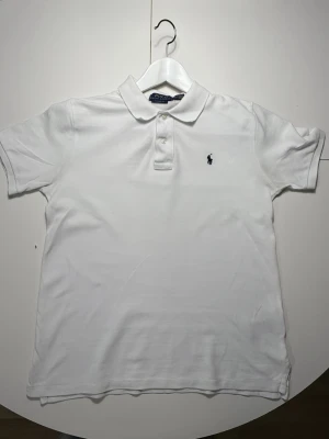 Vit pikétröja från Polo Ralph Lauren - Klassisk vit pikétröja från Polo Ralph Lauren med korta ärmar och krage. Tröjan har knappar vid halsen och den ikoniska broderade loggan på bröstet. Tillverkad i mjuk bomull som känns skön mot huden. Modellen är storlek barn 14-16 år