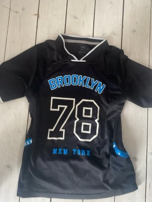 Svart sporttröja Brooklyn 78 - Snygg svart sporttröja med texten BROOKLYN och siffran 78 i vitt och blått framtill. Tröjan har korta ärmar, vit kant vid kragen och blå detaljer på sidorna samt texten NEW YORK. Perfekt för träning eller streetwear.