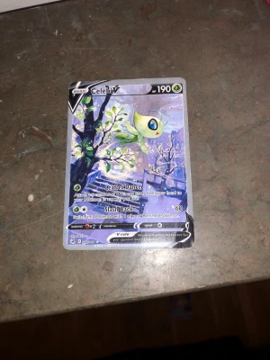 Pokémonkort Celebi V - Säljer ett Pokémonkort av Celebi V med 190 HP. Kortet har en färgstark design med Celebi i ett vinterlandskap och detaljer i grönt, blått och lila. Kortet är gjort av laminerad kartong och har klassisk Pokémon-baksida. Perfekt för samlare eller spelare. Kortet är fejk