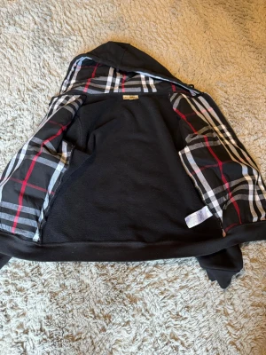 Svart hoodie med rutigt foder - Säljer en svart hoodie från Burberry med dragkedja och klassiskt rutigt foder i huvan och insidan i mycket bra skick. Hoodien har fickor framtill och justerbar huva med snören. Perfekt för dig som gillar stilrena detaljer och vill ha något som sticker ut lite extra.