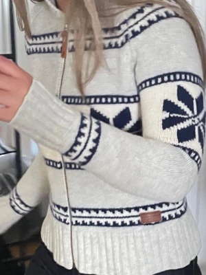 Grå Vit peak performance stickad  kofta med blått mönster - Säljer en grå vit peak performance stickad kofta med dragkedja och blått nordiskt mönster. Koftan har hög krage, långa ärmar och ribbade muddar. Perfekt för kalla dagar och har en snygg retrovibe. Märke syns på läderpatch vid nederkanten.