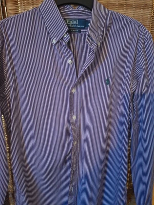 Randig skjorta från Polo Ralph Lauren - Snygg lila och vit randig skjorta från Polo Ralph Lauren i slim fit. Skjortan har klassisk krage, knappar framtill och den ikoniska gröna loggan broderad på bröstet. Tillverkad i bomull och perfekt för en clean och stilren look.