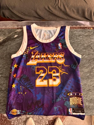 Lakers basketlinne LeBron James #23 - Snyggt basketlinne från Nike med Lakers tryck och nummer 23, inspirerat av LeBron James. Lila och gult med vita kanter, coola grafiska detaljer och NBA-logga. Perfekt för basketträning eller att bära som streetwear.