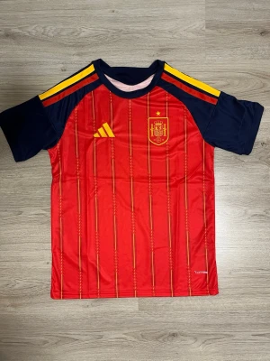Spanien hemmatröja VM perfekt till sommaren - Säljer en officiell röd matchtröja från Spaniens landslag, tillverkad av Adidas. Tröjan har gula detaljer, mörkblå ärmar och axelparti samt tunna vertikala ränder. Materialet är lätt och andas, perfekt för träning eller match.