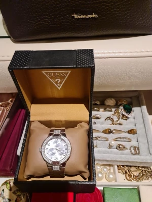 Silverfärgad klocka från Guess - Köpt för 2500kr. Stilren och lyxig klocka från Guess i silverfärg med metallarmband. Urtavlan är också silverfärgad och har tydliga siffror samt tre mindre urtavlor. Boetten är dekorerad med gnistrande stenar för extra bling. Perfekt accessoar för dig som gillar en elegant look.