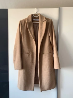 Klassisk beige kappa från Ellos - Stilren beige kappa från ellos med rak passform och diskreta fickor på sidorna. Kappan har klassisk krage. Perfekt för dig som gillar tidlös och enkel design.