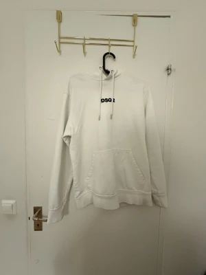 Vit hoodie från DSQ2 med huva - Säljer en stilren vit hoodie från DSQ2 med svart logga på bröstet. Hoodien har en klassisk känguruficka, dragsko i huvan och ribbade muddar. Perfekt för dig som gillar enkel och clean streetwear. Materialet känns mjukt och bekvämt.