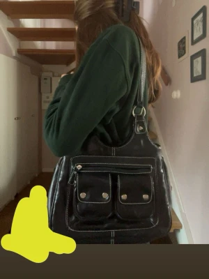 Svart axelväska med fickor - Snygg svart handväska i skinnimitation med två framfickor med knappar och en dragkedjeficka. Väskan har bred axelrem och vita kontrastsömmar som ger en cool look. Perfekt storlek för det viktigaste och enkel att matcha till olika outfits. 💕