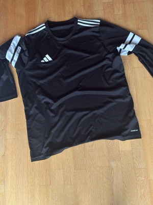 Svart långärmad tröja med vita inslag från Adidas - svart långärmad sporttröja från Adidas. Tröjan är kolsvart, med vita detaljer, bilden inte rättvisAeroready med vita ränder på axlar och ärmar samt det klassiska Adidas-loggan på bröstet. Tillverkad i ett lätt och snabbtorkande funktionsmaterial storlek XXL men mer som XL. Fläck på baksidan se bild 3