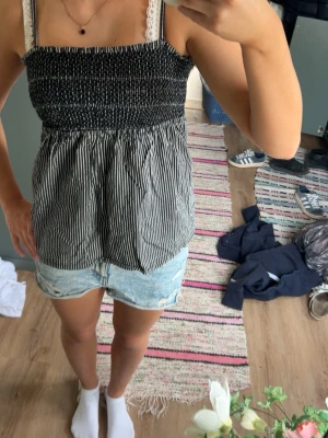 Svartvit randig linnetopp med spets - Söt svartvit linnetopp med smockad överdel och tunna axelband. Toppen har ett randigt mönster nedtill och sitter löst från midjan. Perfekt för varma dagar och enkel att styla med jeansshorts.