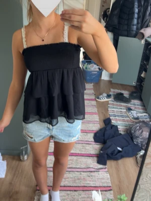 Svart volang topp med spetsband - Säljer en svart topp med volanger och smockad överdel. Toppen är luftig och har en lätt utsvängd passform, perfekt för varma dagar.